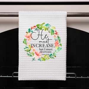 Pu&ograve; includere: Asciugamano da cucina bianco con un design a corona di fiori e il testo "He must increase, but I must decrease. John 3:30".