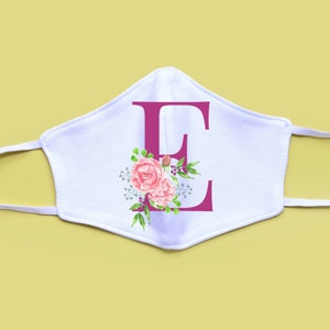 K&ouml;nnte beinhalten: Wei&szlig;e Gesichtsmaske mit einem rosafarbenen Blumendesign und dem Buchstaben "E" in Rosa.