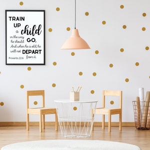 May include: A black and white framed print with the text "Train up a child in the way he should go, And when he is old, he will not depart from it. Proverbs 22:6" hanging on a white wall with gold polka dots. Two wooden chairs and a white table with a wire basket are in the foreground.