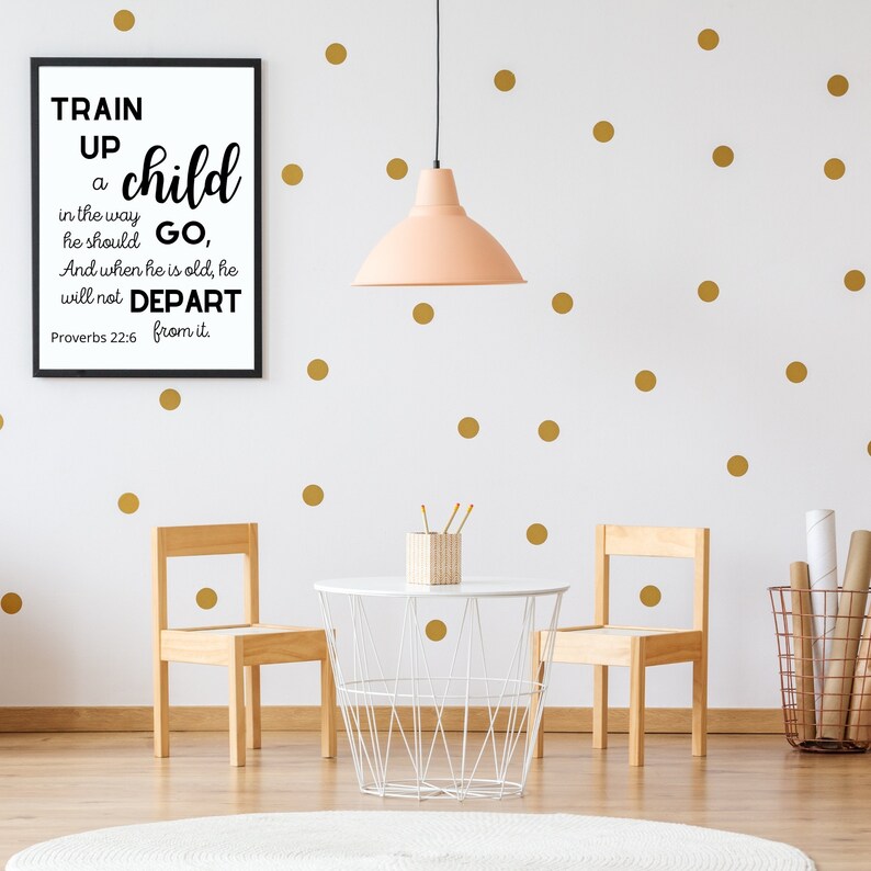 May include: A black and white framed print with the text "Train up a child in the way he should go, And when he is old, he will not depart from it. Proverbs 22:6" hanging on a white wall with gold polka dots. Two wooden chairs and a white table with a wire basket are in the foreground.