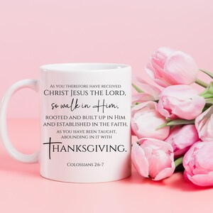 May include: White ceramic mug with a black inspirational quote from Colossians 2:6-7. The quote reads "As you therefore have received Christ Jesus the Lord, so walk in Him, rooted and built up in Him and established in the faith, as you have been taught, abounding in it with Thanksgiving." The mug is sitting on a pink surface with a bouquet of pink tulips.