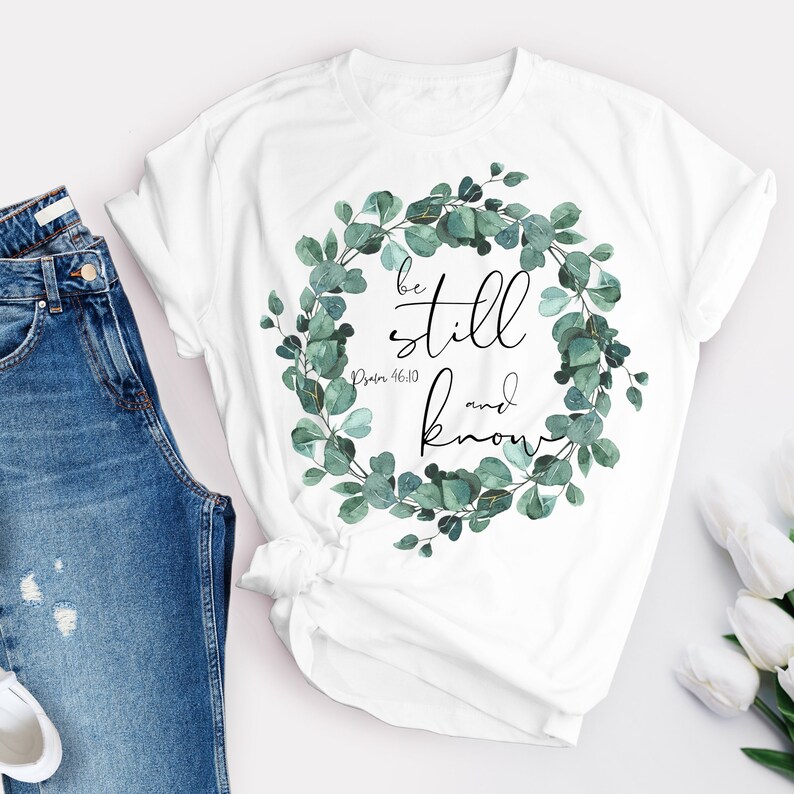 Puede incluir: Camiseta blanca con un dise&ntilde;o de corona floral verde y el texto "be still and know" con la referencia b&iacute;blica "Salmo 46:10".