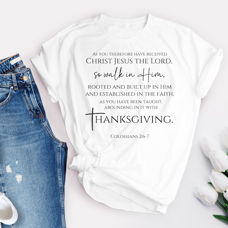 May include: White t-shirt with a black print of a Christian quote about Thanksgiving. The quote reads "As you therefore have received Christ Jesus the Lord, so walk in Him, rooted and built up in Him and established in the faith, as you have been taught, abounding in it with Thanksgiving." The quote is followed by "Colossians 2:6-7".