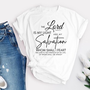 Op de afbeelding: Wit T-shirt met zwarte tekst die luidt: "THE Lord IS MY LIGHT PSALM 27:1 AND MY Salvation WHOM SHALL I FEAR? THE LORD IS THE STRENGTH OF MY LIFE; OF WHOM SHALL I BE AFRAID?"