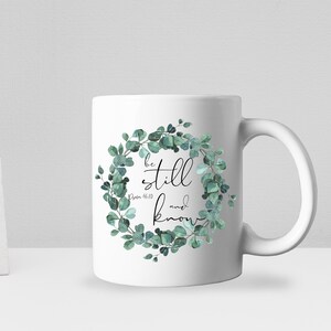 Puede incluir: Taza de cer&aacute;mica blanca con un dise&ntilde;o de corona de eucalipto verde. La corona tiene detalles dorados y el texto "be still and know" con la referencia b&iacute;blica "Salmo 46:10".