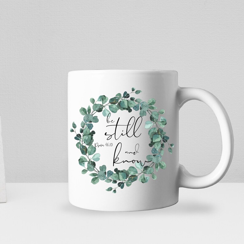 Puede incluir: Taza de cer&aacute;mica blanca con un dise&ntilde;o de corona de eucalipto verde. La corona tiene detalles dorados y el texto "be still and know" con la referencia b&iacute;blica "Salmo 46:10".