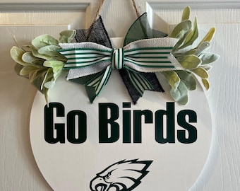 May include: A round white wooden door sign featuring the dark green text Go Birds and a dark green eagle logo. The sign is decorated with a green and white striped burlap bow, black mesh ribbon, and artificial sage green leaves, hanging by twine.
