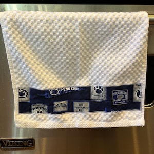 May include: White kitchen towel with a textured pattern and a navy blue band featuring Penn State University logos and text. The towel is hanging on a stainless steel oven.