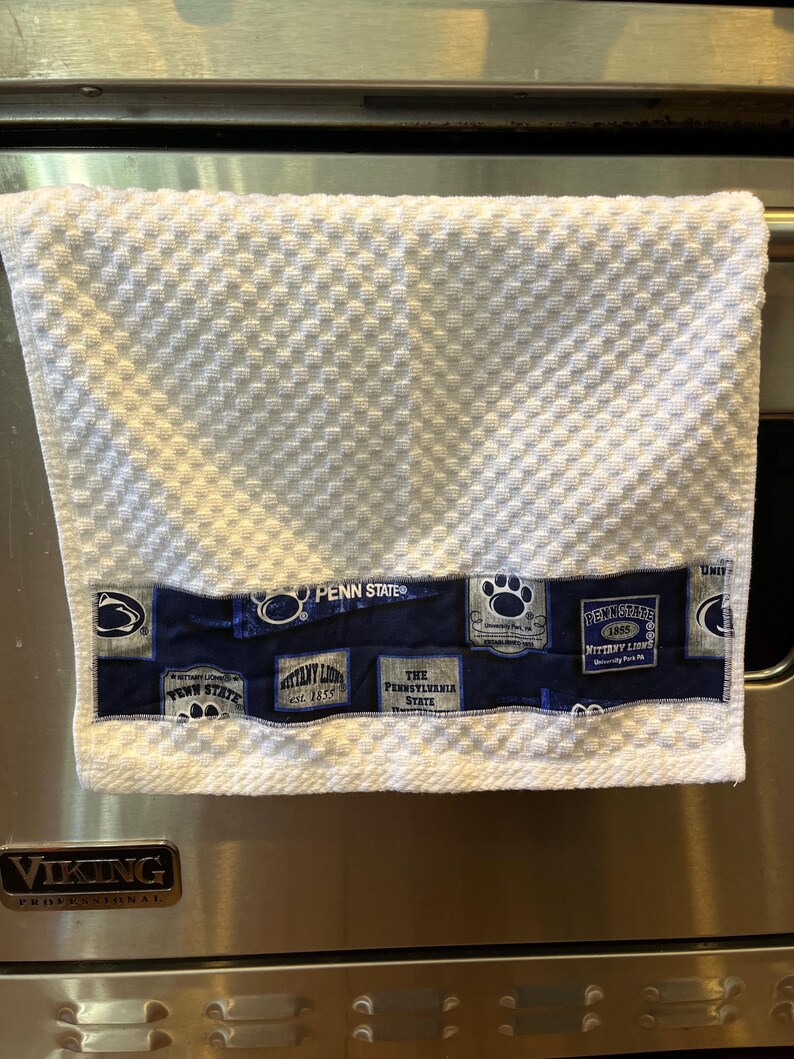 May include: White kitchen towel with a textured pattern and a navy blue band featuring Penn State University logos and text. The towel is hanging on a stainless steel oven.