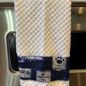 May include: White textured kitchen towel with a navy blue band featuring Penn State logos and text. The towel has a diamond pattern and includes the text "PENN STATE", "Nittany Lions", and "The Pennsylvania State".