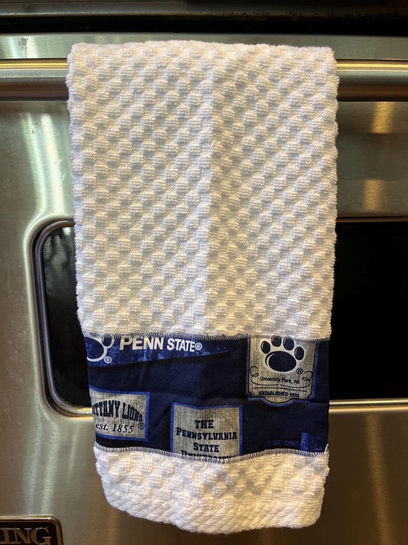 May include: White textured kitchen towel with a navy blue band featuring Penn State logos and text. The towel has a diamond pattern and includes the text "PENN STATE", "Nittany Lions", and "The Pennsylvania State".