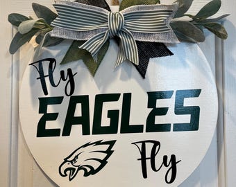 May include: A white round wooden sign with the words "Fly Eagles Fly" in black and green lettering. The sign is decorated with a green and black bow and green leafy sprigs.
