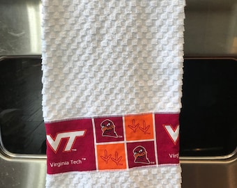 May include: White kitchen towel with a red, orange, and white border featuring the Virginia Tech logo and a Hokie bird. The border also includes the text "Virginia Tech" and "VT".