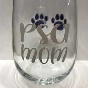 May include: A clear glass wine glass with a white vinyl decal that says "Paw Mom" with two blue paw prints above the text.