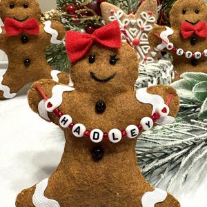 May include: Gingerbread man ornament with a red bow and a beaded necklace spelling "HADLEY". The brown felt ornament has white icing details and black button eyes. Other gingerbread men and holiday greenery are in the background.