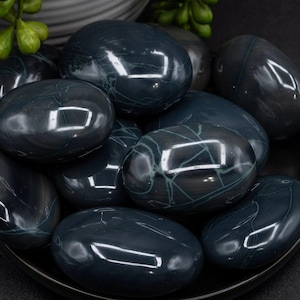 May include: A collection of polished, oval-shaped stones in shades of dark teal and gray, arranged in a black bowl. The stones have a glossy finish with subtle, light green veining. The arrangement is on a dark surface.