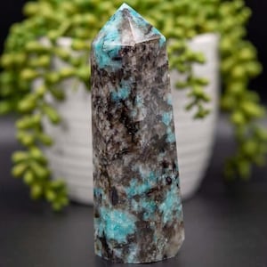 May include: A teal and black amazonite crystal point. The crystal is polished and has a pointed tip. The crystal is about 4 inches tall.