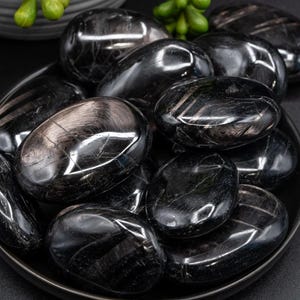 May include: A collection of polished black stones, oval-shaped, displayed in a dark gray bowl. The stones have a glossy, reflective surface with subtle internal patterns. The arrangement suggests a natural, organic theme, possibly for decorative or metaphysical purposes.