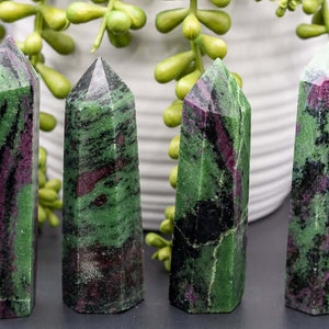 May include: Four green and red zoisite crystal towers with pointed tops. The crystals have a mix of green and red hues, with dark black inclusions. The crystals are arranged in a row, with a blurred background of greenery and a white pot.