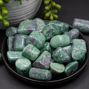 May include: A close-up of a bowl filled with green and pink tumbled ruby zoisite stones. The stones are smooth and polished, with a variety of shapes and sizes.