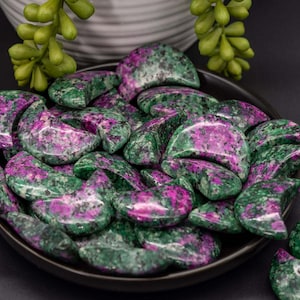 May include: A collection of polished ruby in zoisite crescent-shaped stones, displayed in a dark brown bowl. The stones feature a mix of deep green and vibrant pink hues, with a smooth, glossy finish. The bowl is set against a dark background.