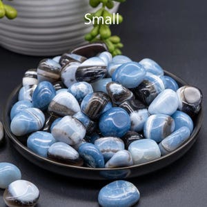 May include: A close-up of a bowl filled with small, blue and white banded agate tumbled stones. The stones are smooth and polished, and the bowl is black. The word "Small" is written in white text above the bowl.