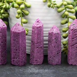 May include: Six purple crystal points with a rough, textured surface. The points are arranged in a row on a black surface.