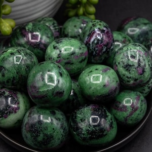 May include: A collection of polished, spherical Zoisite crystals with Ruby inclusions. The stones are primarily green with dark red and black accents. They are arranged in a dark bowl, with a few green plants in the background.