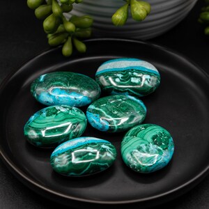 May include: Six polished malachite stones with swirling green and blue patterns rest on a black plate. The stones are oval-shaped and have a glossy finish. A sprig of green foliage is in the background.
