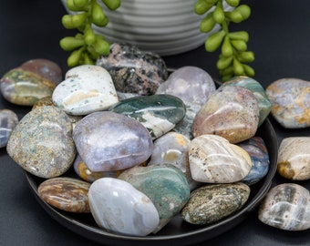 May include: A collection of heart-shaped polished stones in various shades of brown, green, white, and gray. The stones are arranged in a bowl on a black surface.