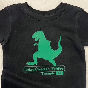 May include: Black t-shirt with a green dinosaur graphic and the text "Token Creature - Toddler  Trample 1/1".