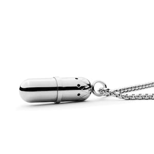 May include: A silver pill-shaped pendant necklace with a chain. The pendant has a detailed design and a small opening at the top.