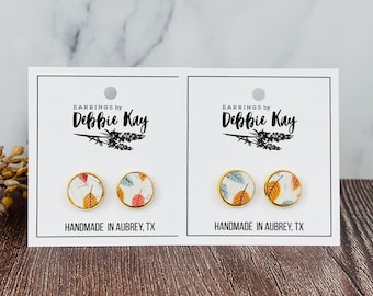 May include: Two pairs of gold-rimmed stud earrings with leaf patterns. The earrings are displayed on white cards with the text "EARRINGS by Debbie Kay" and "HANDMADE IN AUBREY, TX". The earrings feature orange, red, and blue leaves.