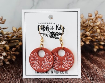 May include: A pair of terracotta-colored, circular earrings with white floral designs and gold-colored hooks. The earrings are displayed on a white card with the text "Earrings by Debbie Kay" and "Handmade in Aubrey, TX."