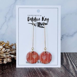 May include: A pair of earrings with a circular, rust-colored design featuring a white arrow pattern. The earrings hang from long, thin gold-colored wires. The card behind the earrings reads "EARNINGS by Debbie Kay".