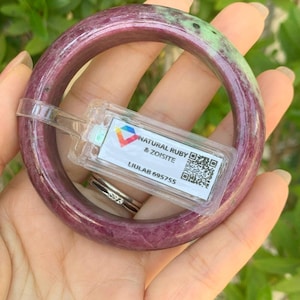 May include: A natural ruby and zoisite bangle bracelet. The bracelet is a deep burgundy color with green accents. The bracelet has a clear tag attached with the text "NATURAL RUBY & ZOISITE" and the code "LIULAB 695755".