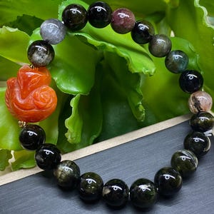 May include: A black and green gemstone bracelet with a red and orange charm. The bracelet is made of round beads and has a gold clasp.