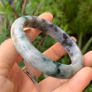 May include: A jade bangle bracelet with a marbled appearance. The bracelet features a mix of white, gray, and green hues. The smooth, polished surface reflects light, highlighting the natural variations in the stone. The bangle is a circular shape.