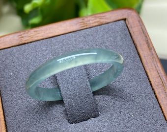 May include: A translucent, pale green jadeite bangle bracelet. The smooth, polished ring is displayed on a gray velvet surface within a wooden box. The jadeite has a subtle, natural color and a glossy finish.