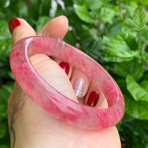May include: A translucent pink quartz bangle bracelet. The bracelet has a marbled appearance with varying shades of pink. The bracelet is held against a background of green foliage.