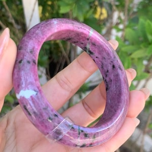 May include: A polished, oval-shaped bangle bracelet in shades of pink and purple, with dark green and black speckles. The bracelet is held in a hand, showcasing its smooth, glossy surface. The background features green foliage.