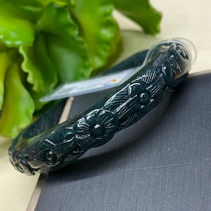 May include: Dark green jade bangle bracelet with carved floral design. The bracelet is a deep forest green color and features detailed carvings of flowers around the entire band. The background includes green foliage and a dark surface.