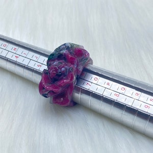 May include: A carved gemstone ring in shades of red, green, and white is displayed on a silver ring sizer. The ring features a detailed floral or organic design. The background is a soft, white, textured surface.
