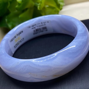 May include: A light blue agate bangle bracelet. The bracelet has a smooth, polished surface with a marbled appearance. The text "Natural Agate" and other details are visible on a sticker attached to the bracelet.