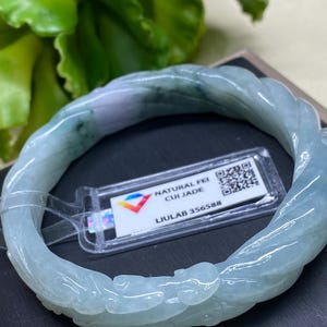May include: A light blue jade bracelet with a carved design. The bracelet has a twisted rope-like pattern and is labeled "NATURAL FEI CUI JADE" with the number "LIULAB 356588". The bracelet is a light blue color with some green and white accents.