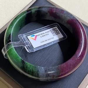 May include: A green, black, and purple jade bangle bracelet with a small tag attached that reads "NATURAL RUBY & MATRIX LIULAB 399769".