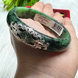 May include: A green jade bangle bracelet with a silver filigree design. The bracelet has a tag attached that reads "TT Nghiên Cứu Kim Cương & Da Quý Tin VDG117621".