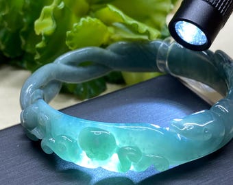 May include: A translucent jadeite bangle bracelet in shades of teal and green. The bracelet features carved aquatic animal designs. A black flashlight shines light on the bracelet, highlighting its details. The background includes green foliage.