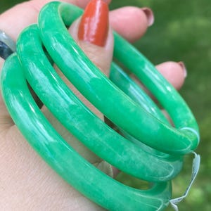May include: Bright green jade bangles. The smooth, polished bracelets are a vibrant shade of green with subtle variations in color. The bangles are stacked together, showcasing their circular design and glossy finish.