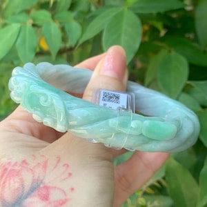 May include: A light green jade bracelet with carved floral and swirling designs. The bracelet is a pale green color and appears to be made of a translucent stone. The bracelet is held in front of a background of green leaves.
